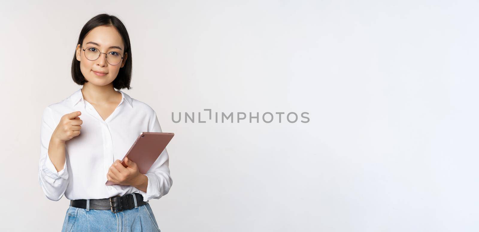 Korean woman, office worker manager in glasses, holding working tablet and pointing at you, choosing recruiting, standing over white background.