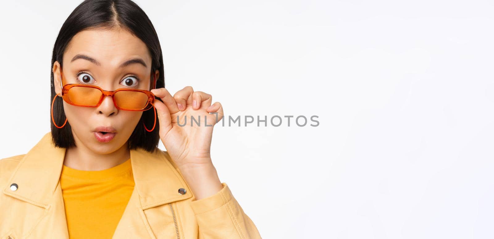 Close up portrait of asian girl looking surprised, wow face, takes off sunglasses and staring impressed at camera, standing over white background.