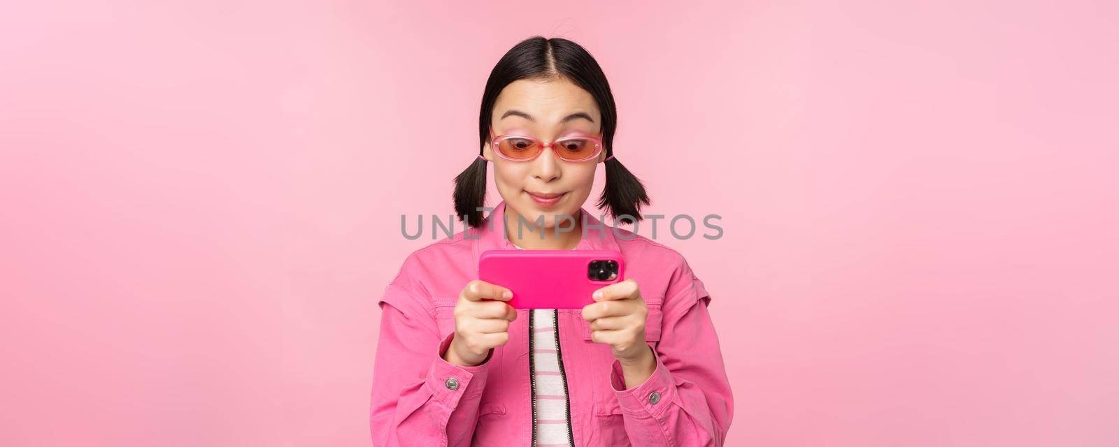 Portrait of happy asian girl playing on smartphone, watching videos on mobile phone app, standing over pink background.