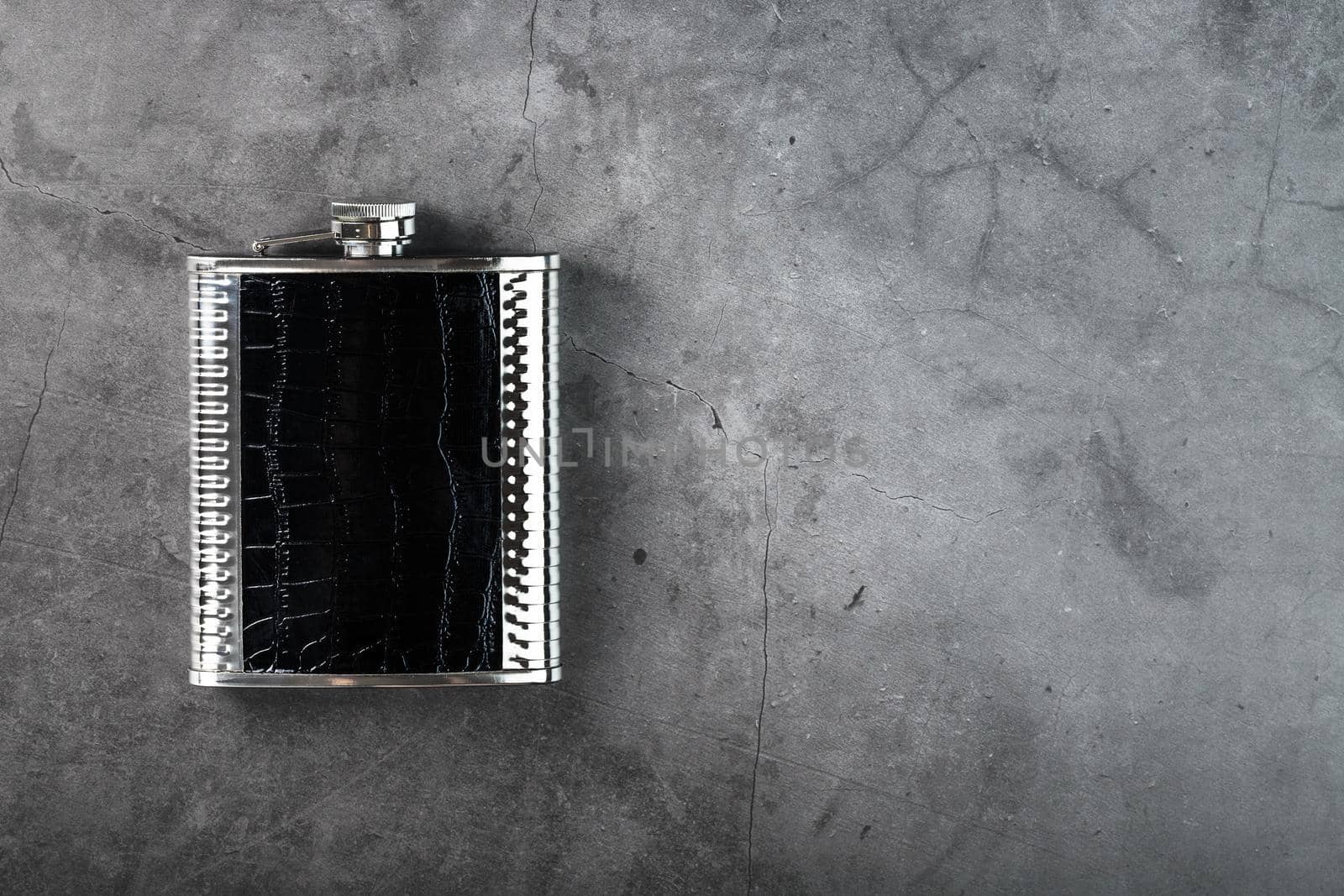 Pocket flask made of steel in leather finish with an alcoholic drink on a dark background with copy space