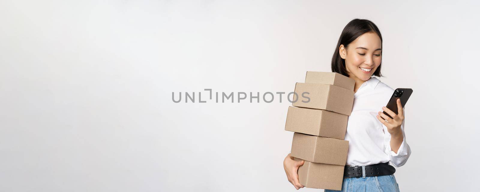Image of young asian woman holding boxes, customer orders and looking at mobile phone, standing over white background.