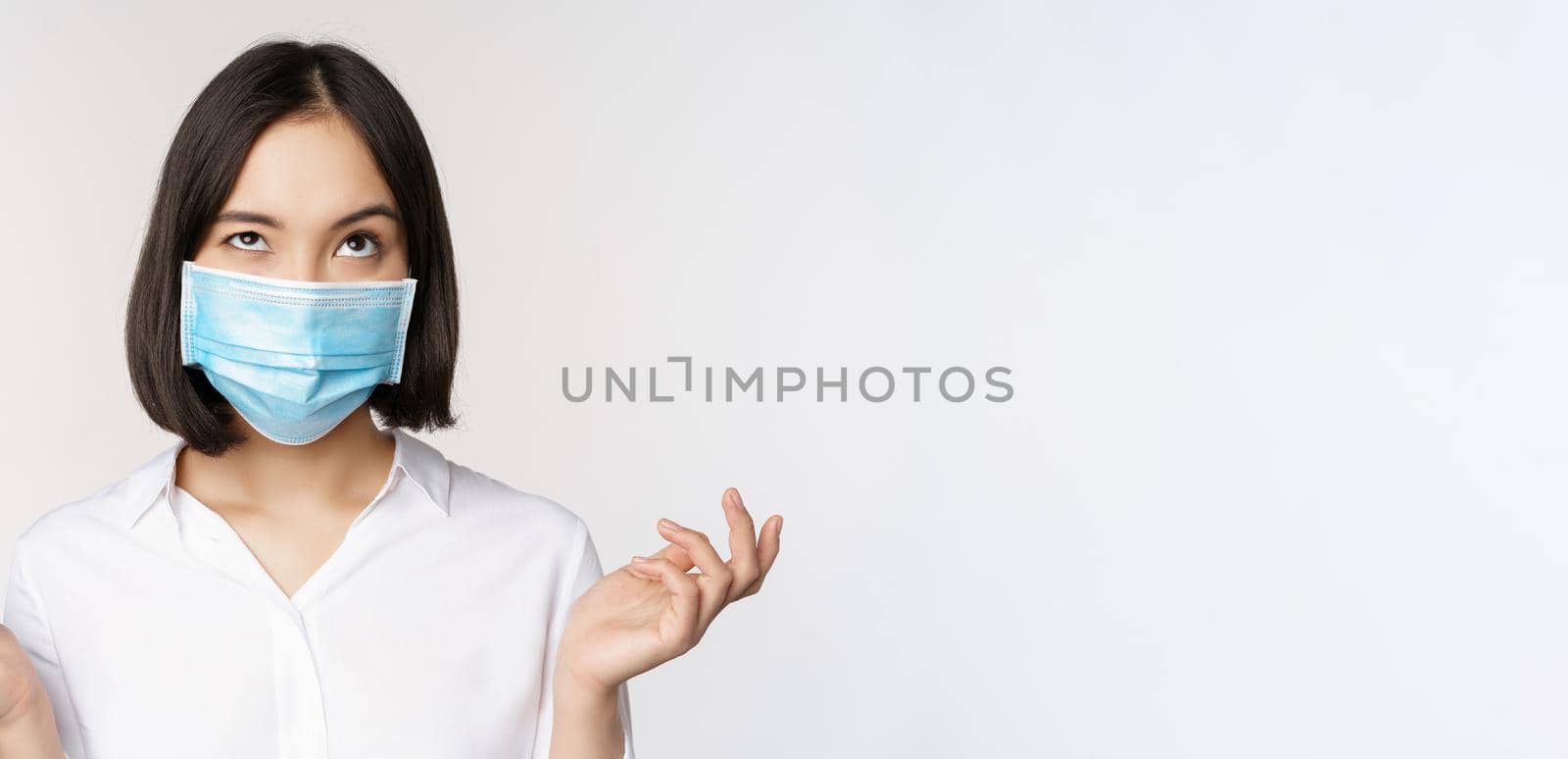 Annoyed asian woman in medical face mask, shrugging and looking up with bothered face expression, standing over white background.