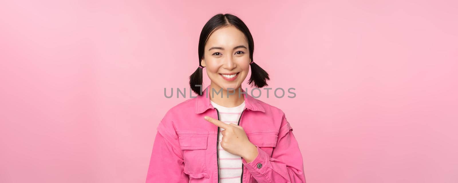 Portrait of beautiful smiling asian girl, pointing finger left, showing advertisement, banner promo, standing over pink background.