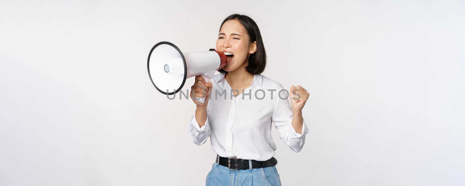 Enthusiastic asian woman, girl activist shouting at protest, using megaphone, looking confident, talking in loudspeaker, protesting, standing over white background.