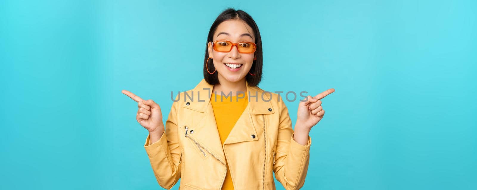 Happy stylish chinese girl in sunglasses, points fingers sideways, invites to check out, see store discount, shows left and right, stands over blue background.