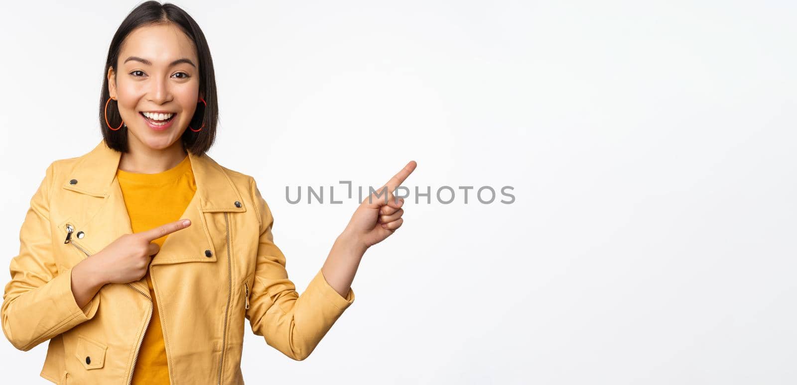 Enthusiastic asian girl pointing fingers right, showing advertisement and smiling, inviting to the store, standing over white background.