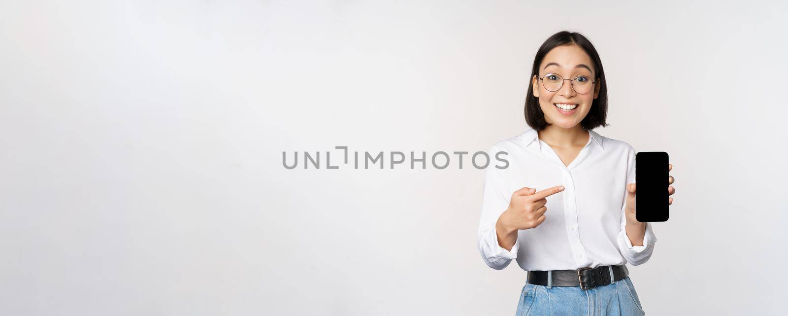 Enthusiastic young asian woman pointing finger at smartphone screen, showing advertisement on mobile phone, white background by Benzoix