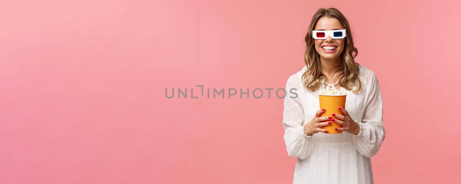 Leisure, going-out and spring concept. Portrait of happy carefree blond girl in white dress enjoying watching movie in 3d, wearing glasses eating popcorn and smiling, attend cinema, pink background by Benzoix