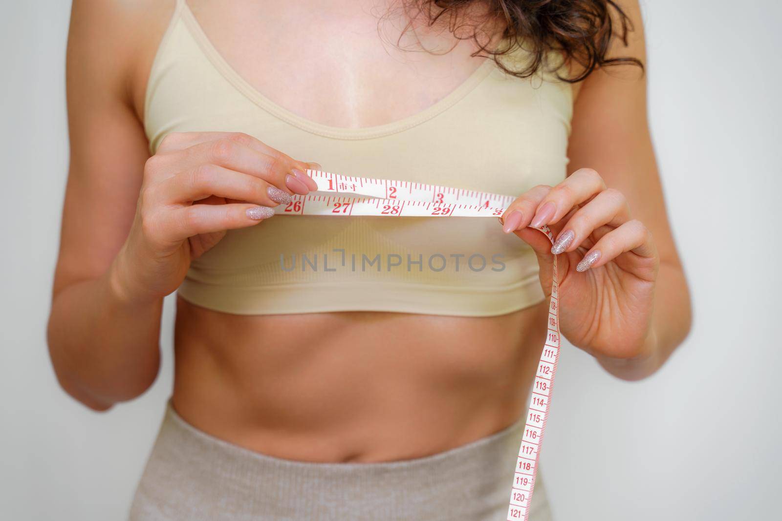 Cropped view of slim woman measuring breasts with tape measure at home, close up. Unrecognizable European woman checks the result of a weight loss diet or liposuction indoors. Healthy lifestyle..