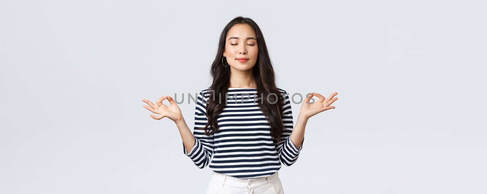 Lifestyle, people emotions and casual concept. Relaxed and patient smiling young asian woman with closed eyes meditating to calm down, do breathing exercises with hands in zen gesture.
