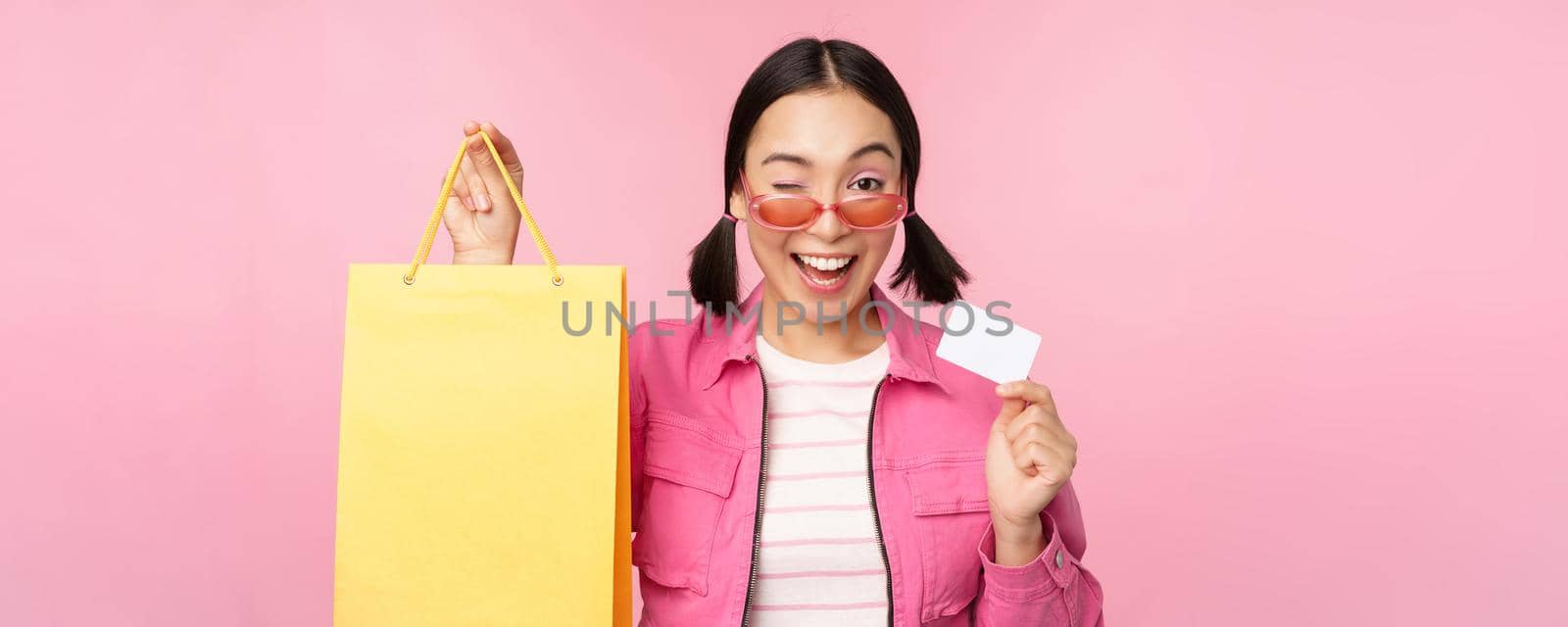 Happy young asian woman showing credit card for shopping, holding bag, buying on sale, going to the shop, store, standing over pink background by Benzoix