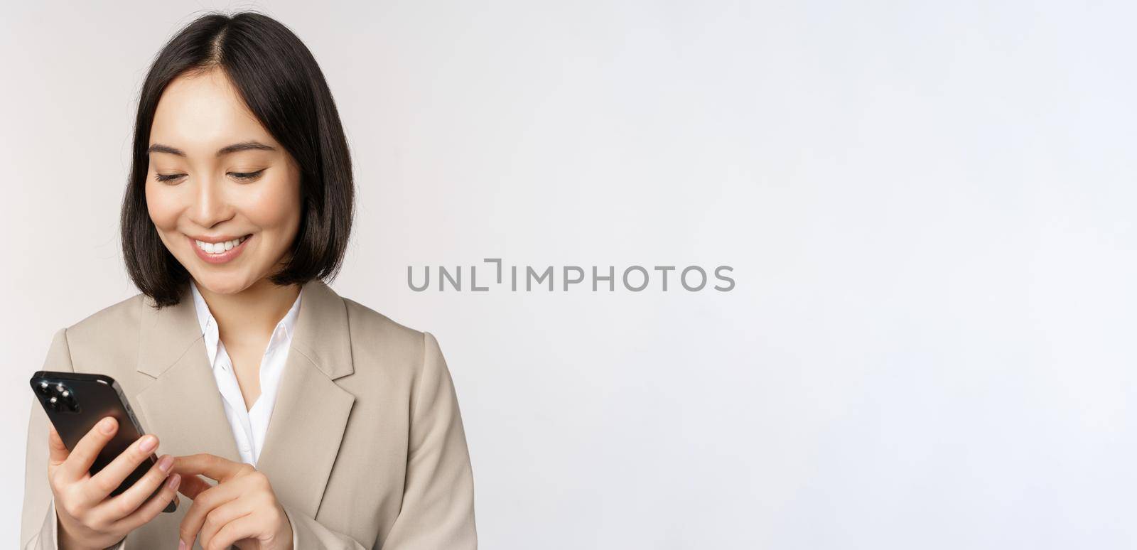 Close up portrait of korean woman, corporate lady in suit, using mobile phone and smiling, holding smartphone, standing over white background.