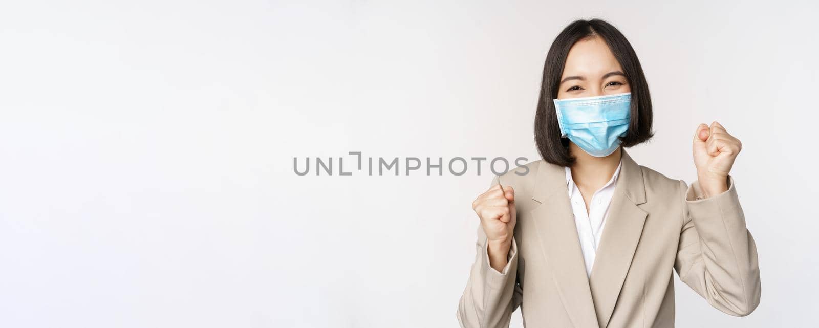 Coronavirus and business people concept. Happy businesswoman in medical face mask dancing, celebrating success, achievement, standing over white background.