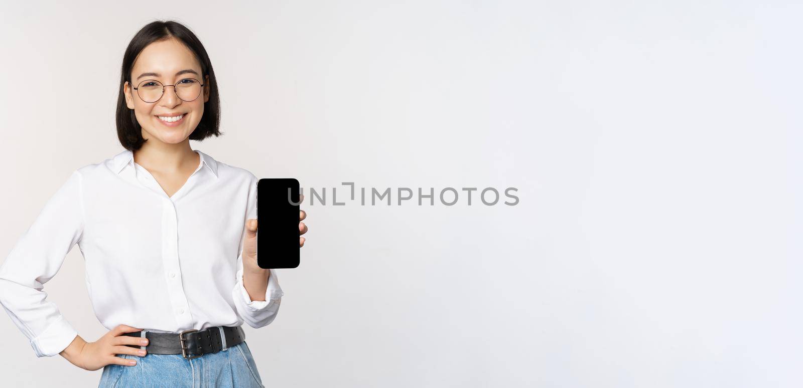 Smiling korean woman showing smartphone screen, demonstrating mobile application, standing over white background.