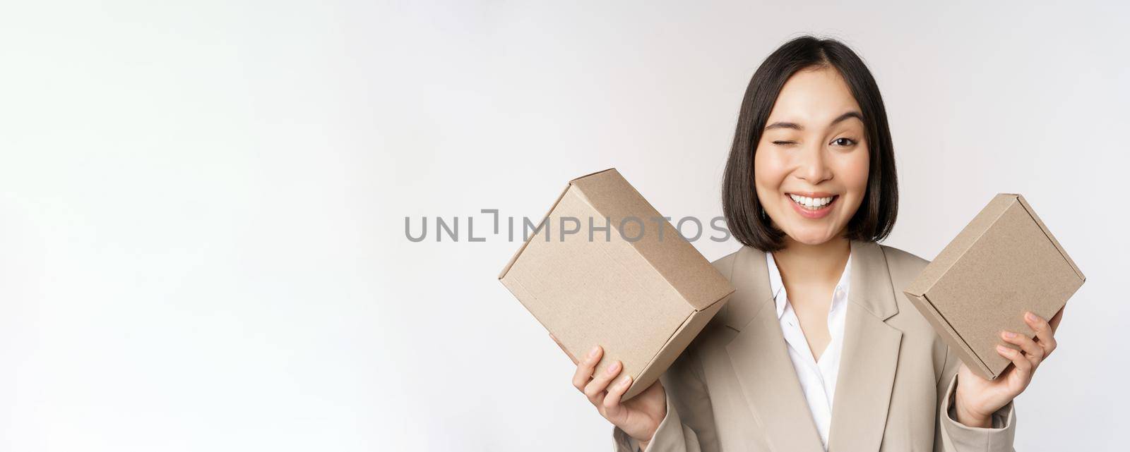 Image of saleswoman, asian businesswoman holding boxes with company brand product, smiling at camera, standing against white background.