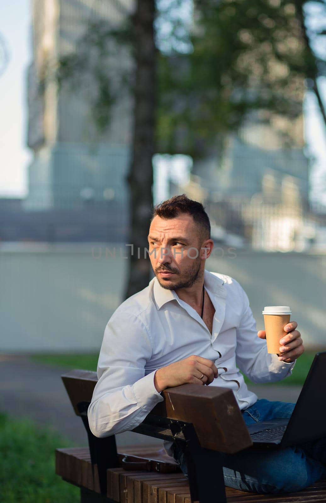 Thoughtful man in the park with a laptop drinking coffee. A young man on a background of green trees, a hot sunny summer day. Warm soft light, close-up.