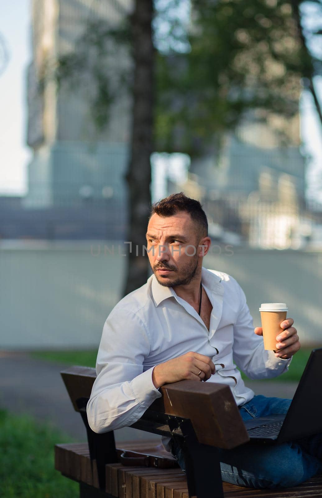 Thoughtful man in the park with a laptop drinking coffee. A young man on a background of green trees, a hot sunny summer day. Warm soft light, close-up.