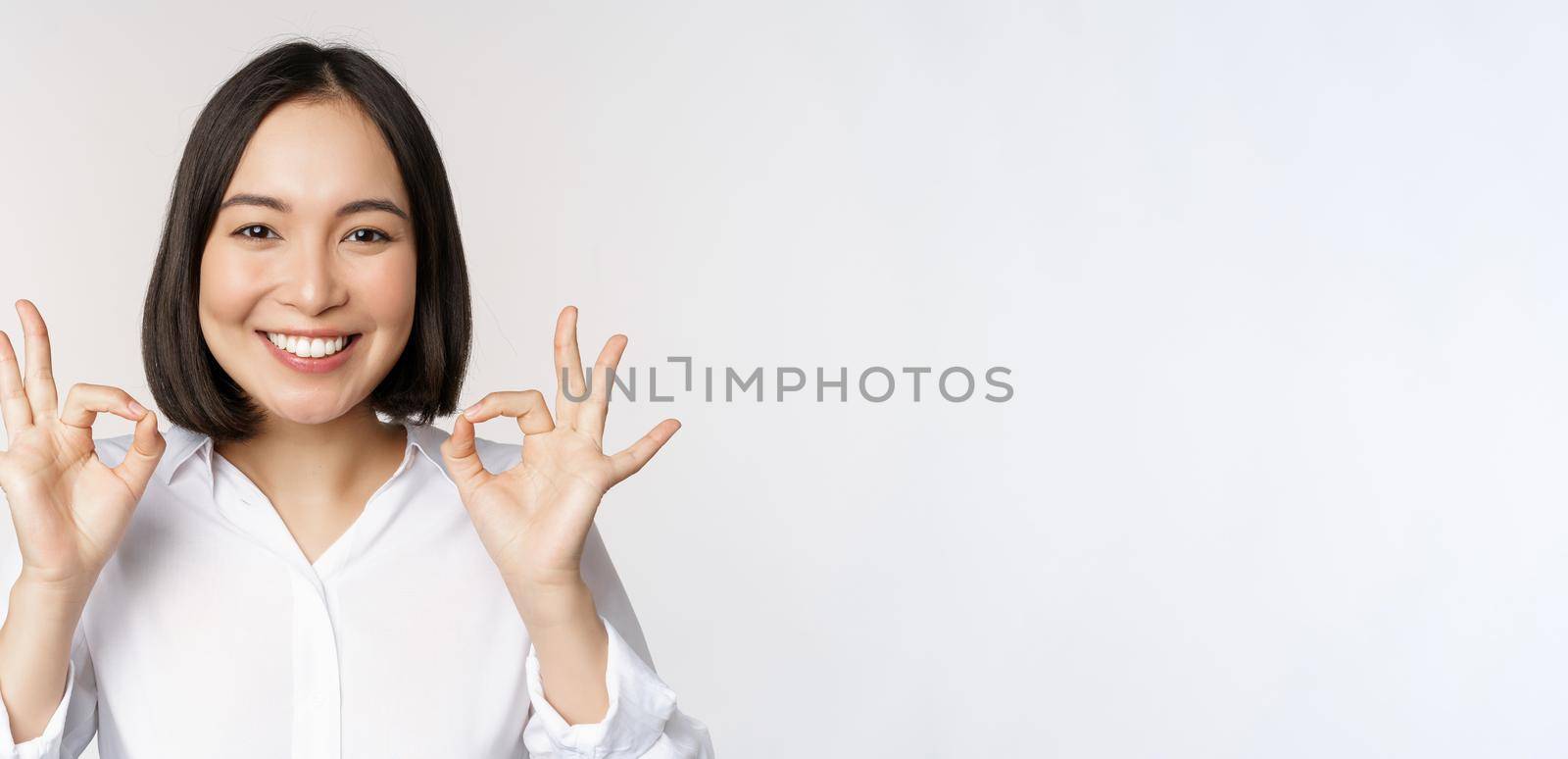 Close up head portrait of asian girl showing okay, ok sign and smiling satisfied, recommending, being pleased, praise and make compliment, white background by Benzoix