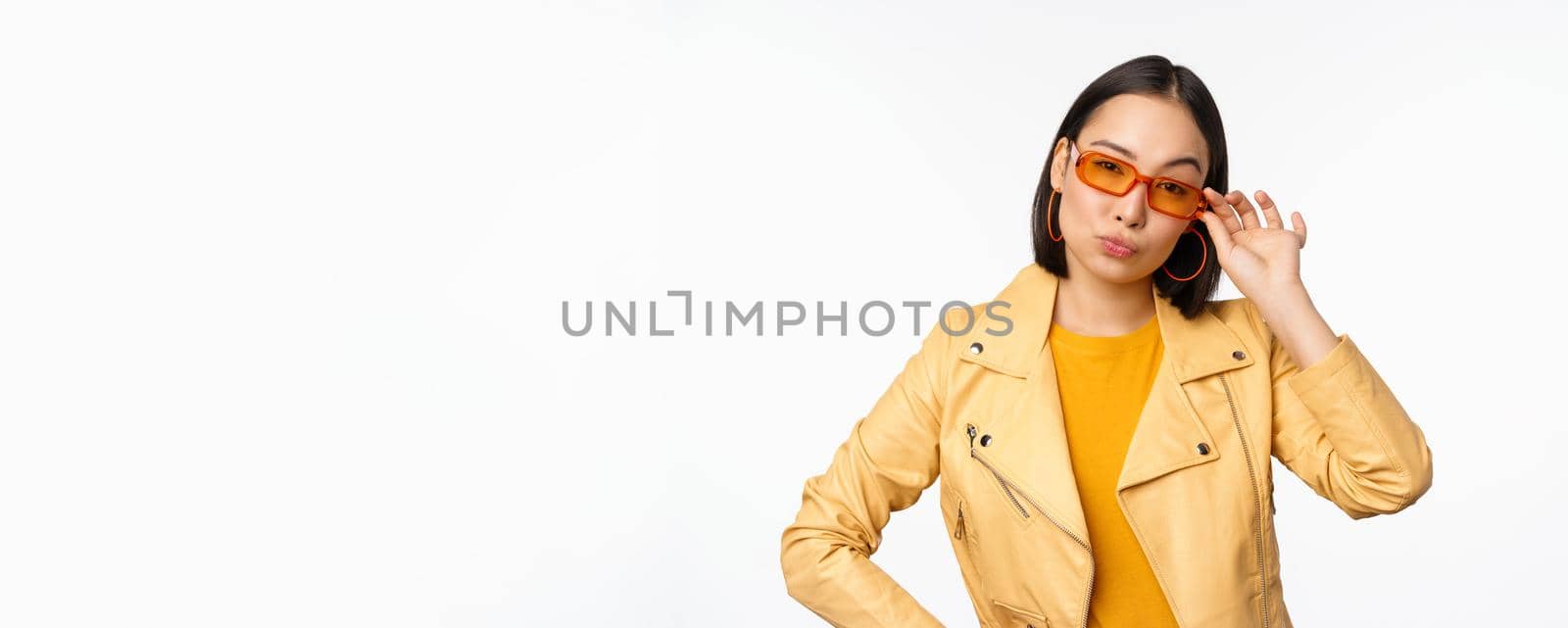 Stylish asian female model, wears sunglasses, looks confident and cool, stands over white background.