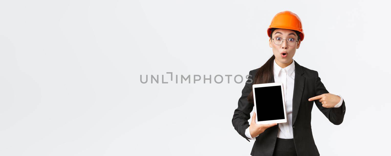 Impressed and surprised asian female engineer, technician at industrial area wear safety helmet and business suit, pointing finger at profit chart, diagram at digital tablet screen, look amazed by Benzoix