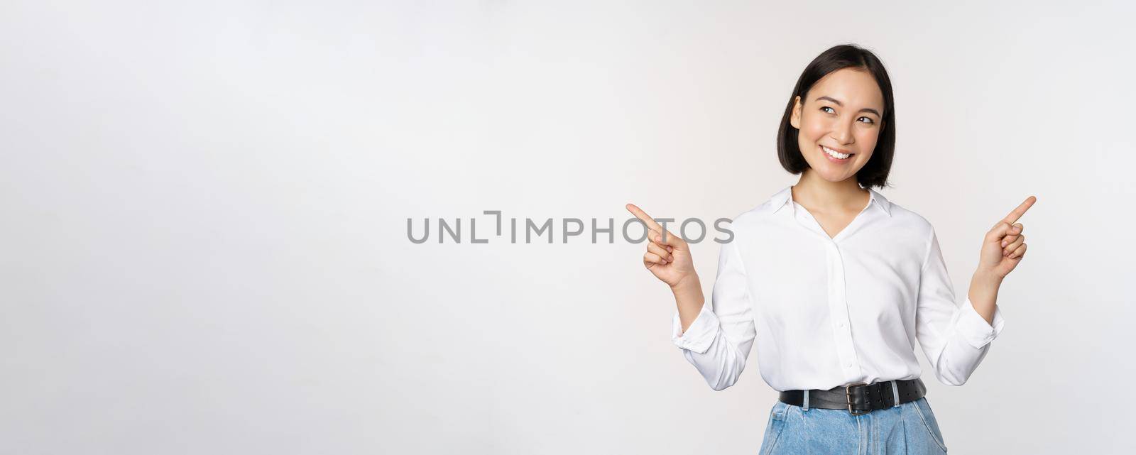 Image of beautiful asian woman pointing fingers left and right, making decision, showing two variants choices, standing over white background.