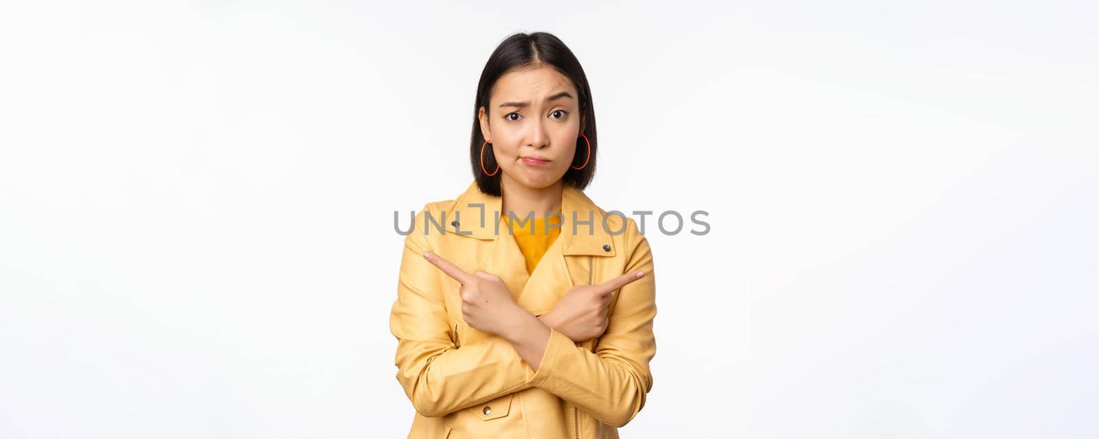 Image of indecisive asian girl, pointing fingers sideways, pointing left and right, choosing variant, deciding, standing over white background. Copy space