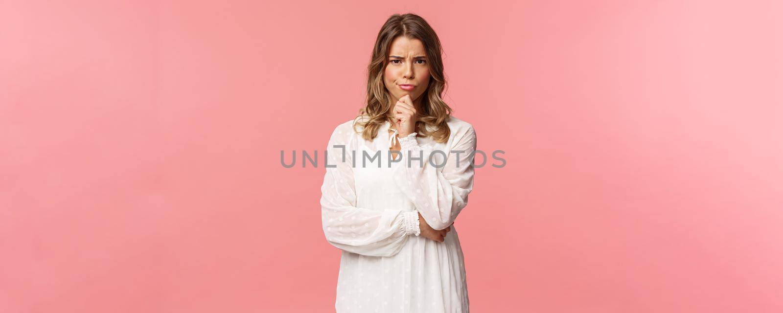 Beauty, fashion and women concept. Portrait of skeptical and judgemental serious-looking blond female in white spring dress, look disbelief, grimacing and frowning, pink background by Benzoix