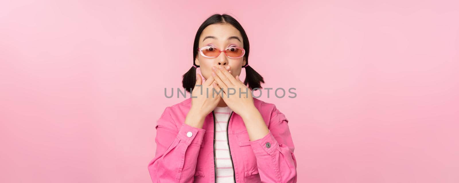 Portrait of shocked, stylish asian girl in sunglasses, closes mouth, looks with surprised face expression, stands over pink background by Benzoix