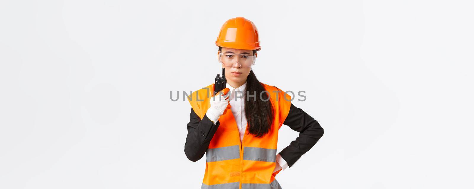 Disappointed asian female construction engineer, technician or industrial manager in safety uniform calling employee via walkie-talkie, scolding personal using radio communication at enterprise.