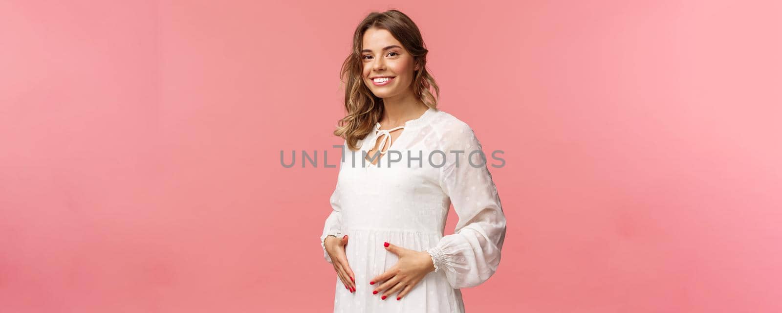 Maternity, women and beauty concept. Tender, cute smiling blond woman in white dress feeling happiness and love, touching belly as expecting child, being pregnant, pink background.