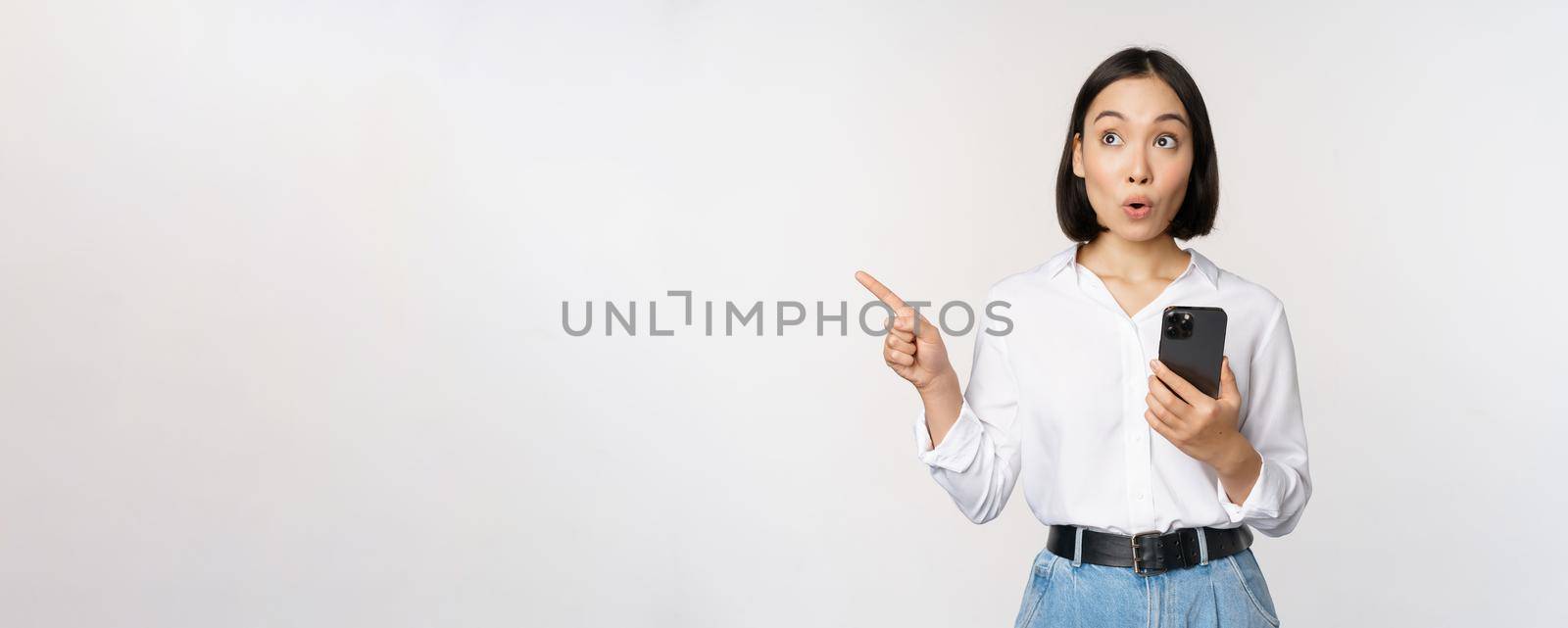 Excited asian girl shopping on mobile app, holding phone and pointing finger left at empty copy space, showing promo text, standing with smartphone against white background by Benzoix