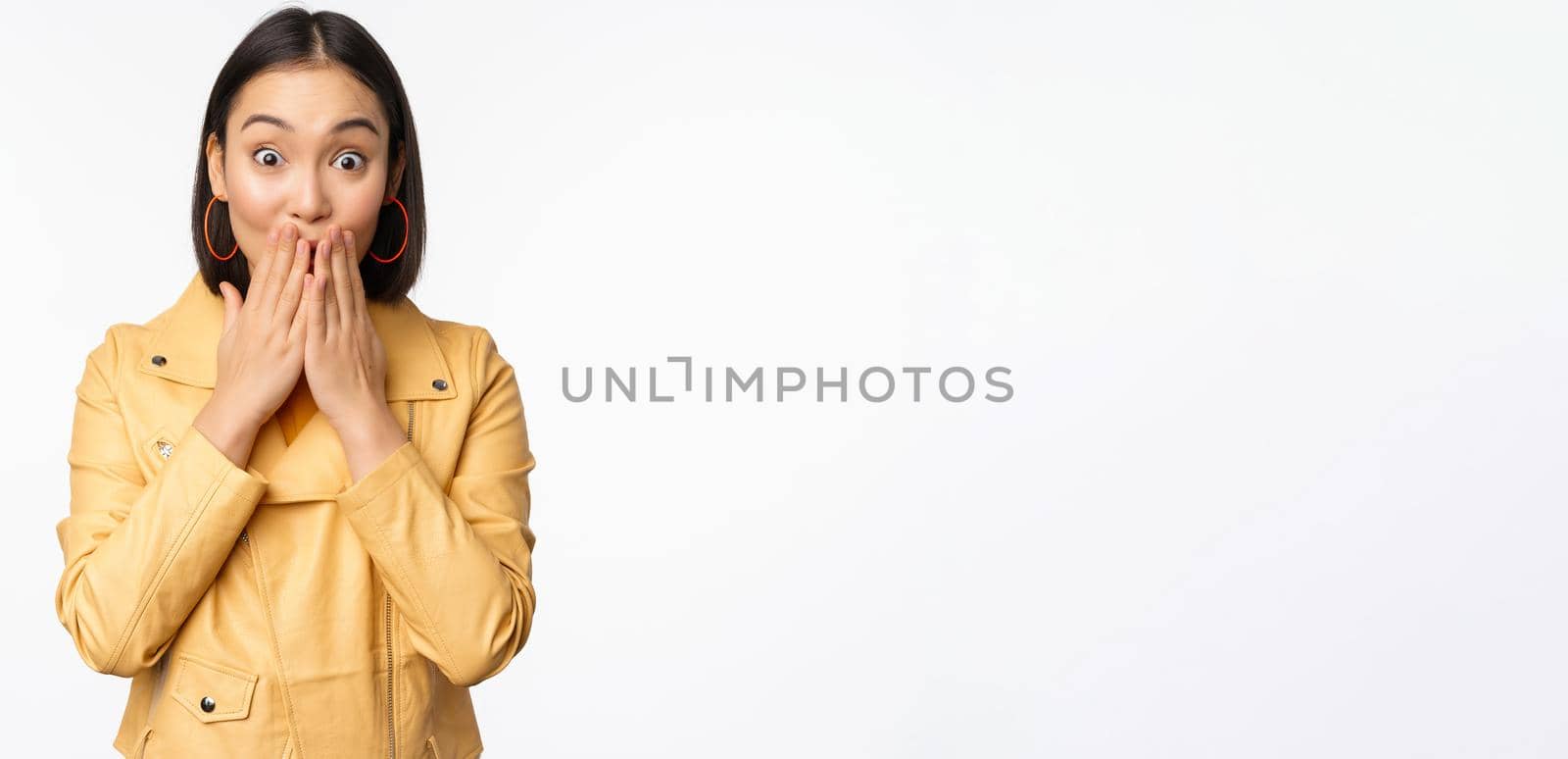Portrait of excited asian girl looking with interest at camera, amazed reaction at big news or announcement, standing in yellow jacket over white background.