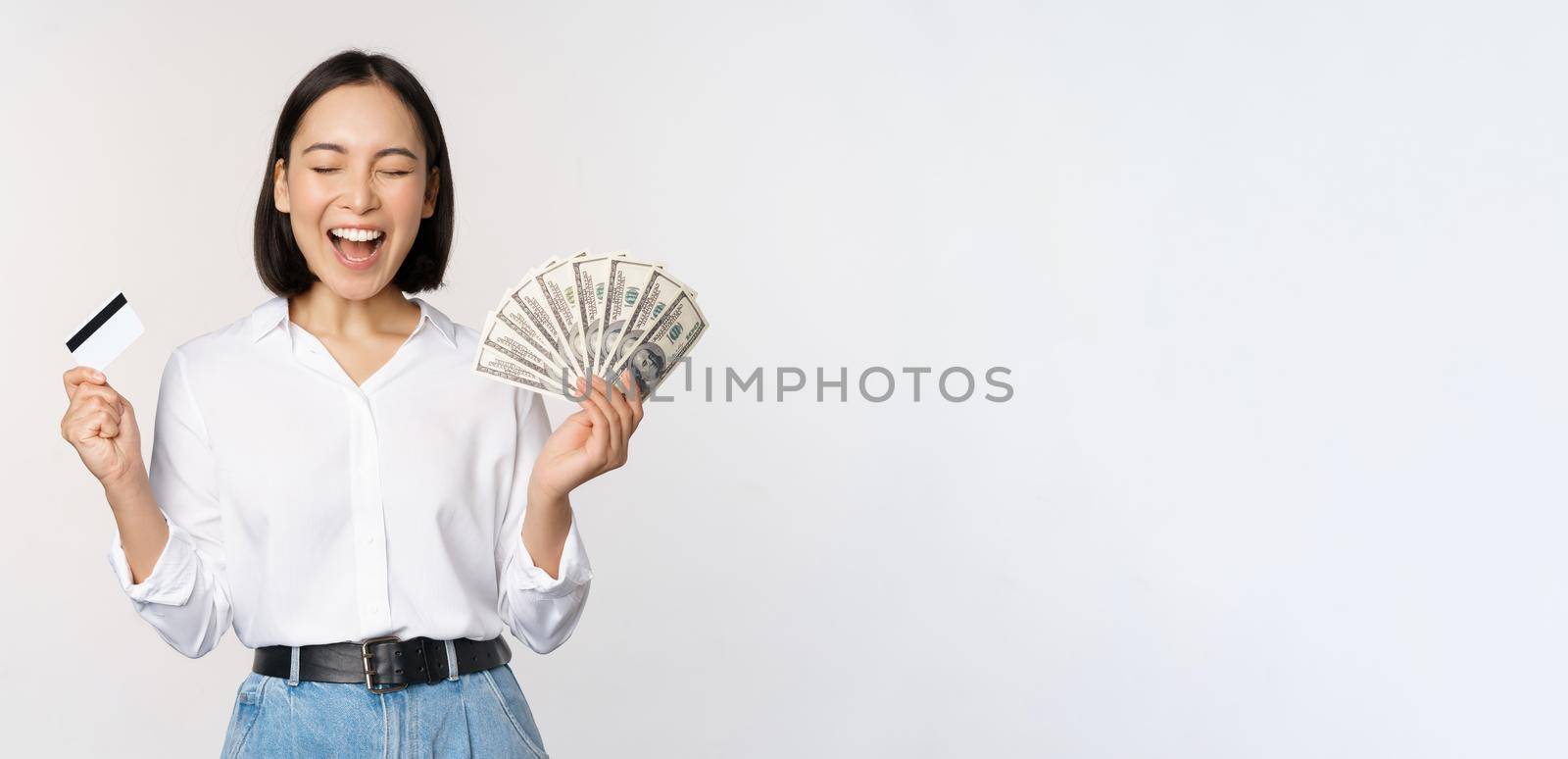 Finance and money concept. Happy young asian woman dancing with cash and credit card, smiling pleased, posing against white studio background by Benzoix