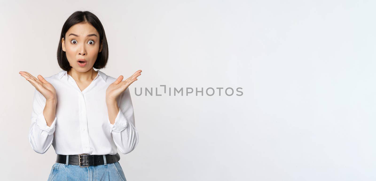 Image of asian female model looking surprised, staring amazed, reacting at surprise big news, standing over white background.