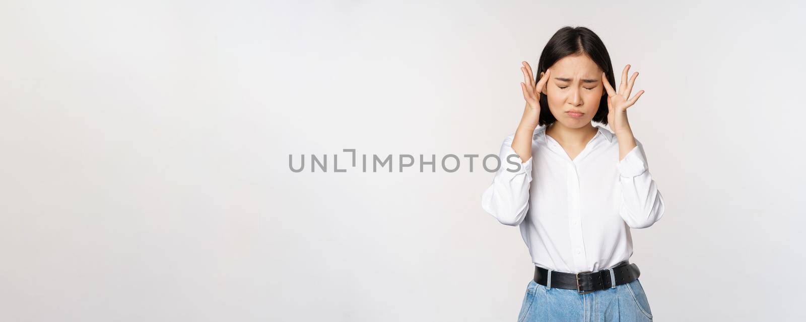 Image of distressed young asian businesswoman, female lady touching head, rubbing temples, has migraine, standing over white background.