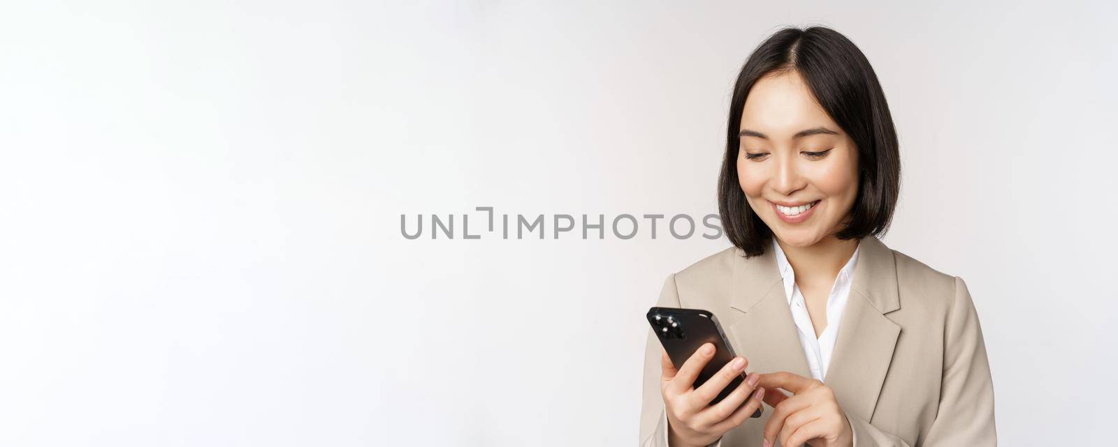 Close up portrait of korean woman, corporate lady in suit, using mobile phone and smiling, holding smartphone, standing over white background by Benzoix