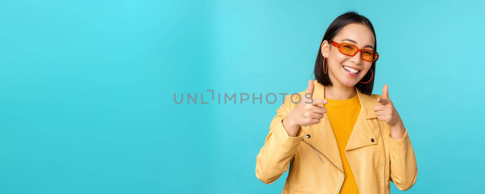 Happy young stylish chinese girl in sunglasses, points fingers at camera with pleased smile, choosing you, congratulating, standing over blue background.