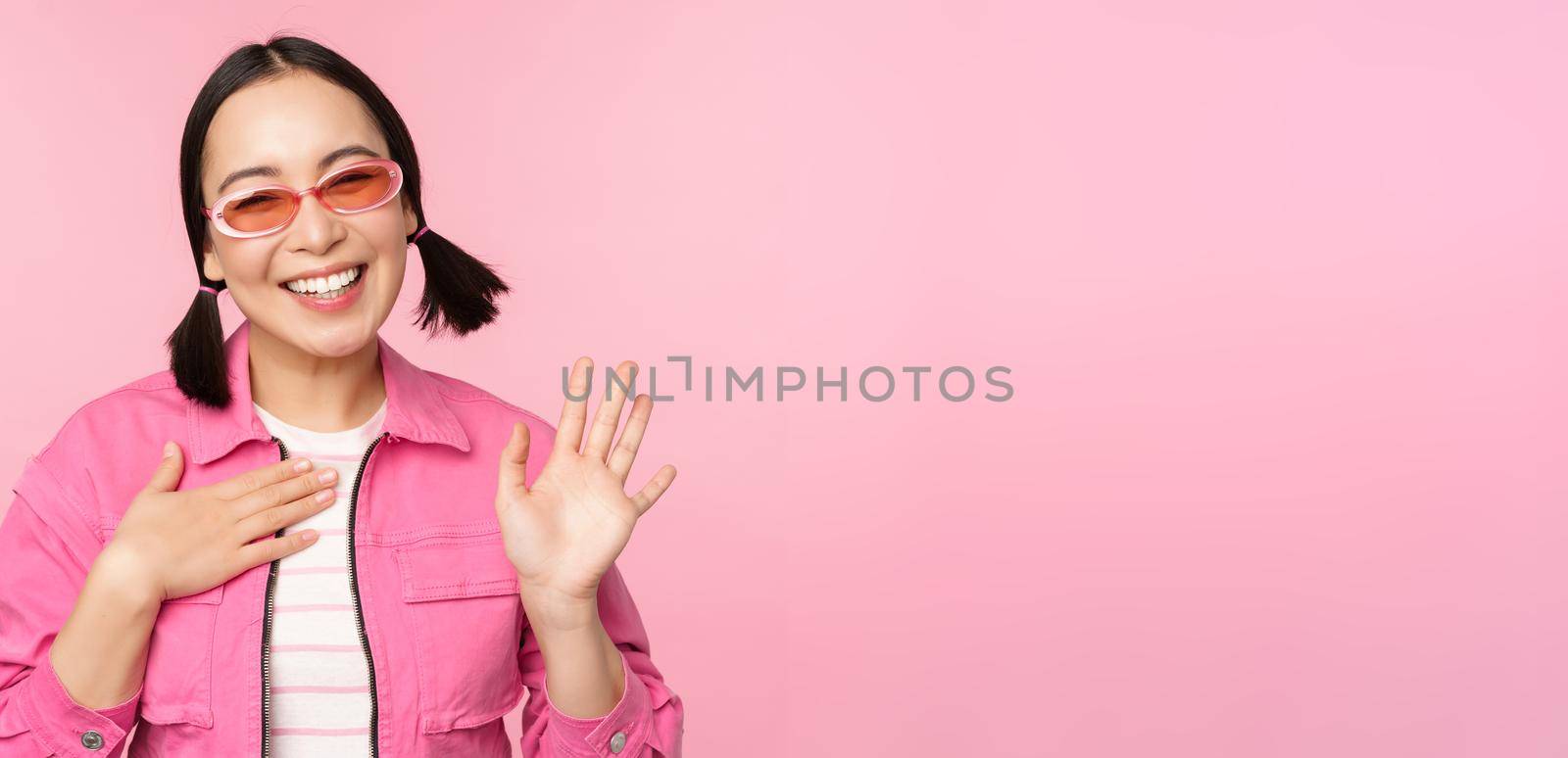 Portrait of friendly asian teen girl in sunglasses saying hello, waves her hand and smiles, greets you, hi gesture, stands over pink background.