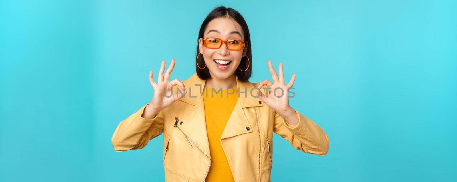 Stylish beautiful asian woman in sunglasses, smiling amazed, showing okay, ok sign, recommending smth, supports excellent choice, standing over blue background.