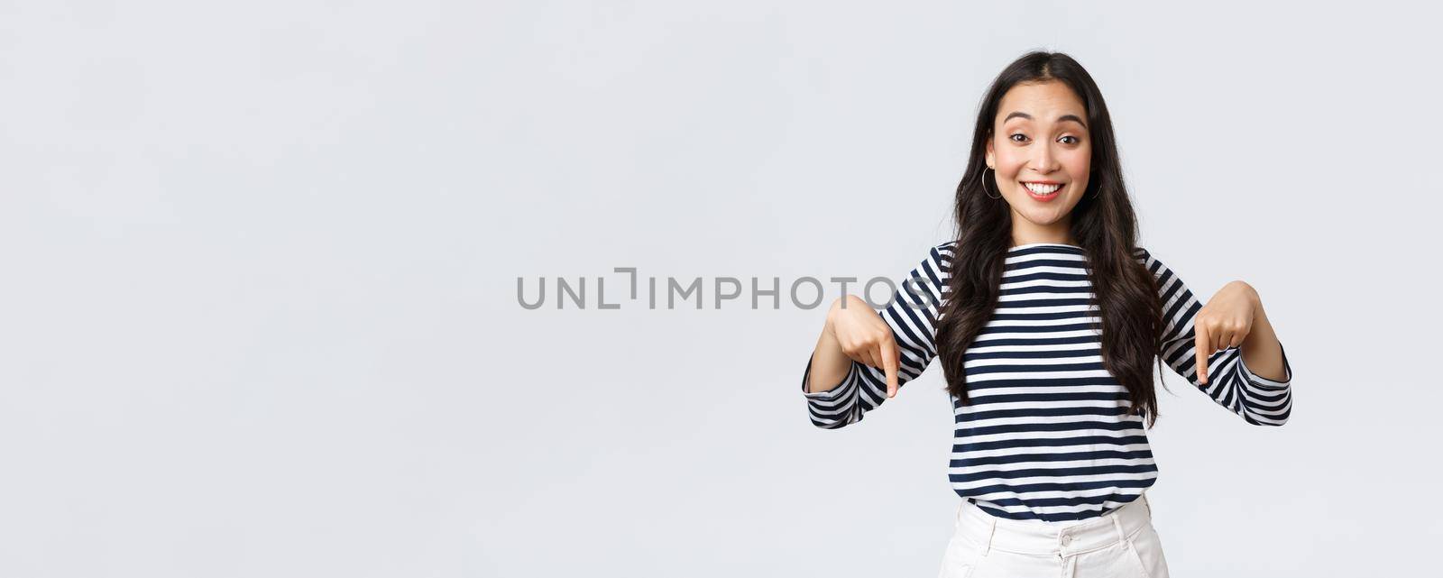 Lifestyle, beauty and fashion, people emotions concept. Excited tender charming asian girl with cute smile pointing fingers down, inviting customers to special event, white background by Benzoix