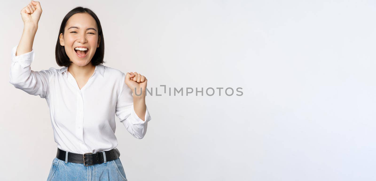 Image of happy lucky asian woman hooray gesture, winning and celebrating, triumphing, raising hadns up and laughing, standing over white background.