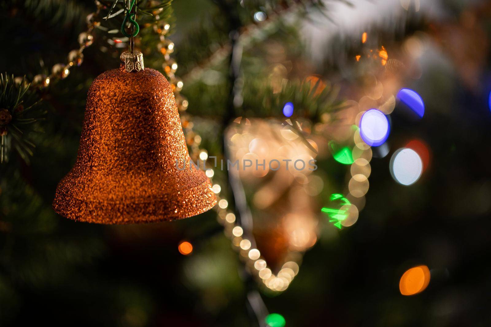 A shiny bell-shaped ornament attached to a Christmas tree in the background with cortical lights.