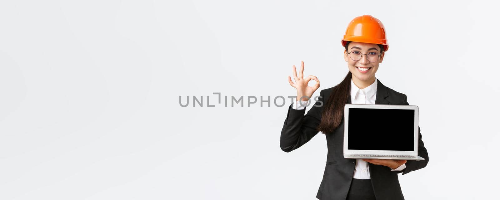 Smiling professional female architect, construction manager at factory showing graph, positive diagram, make okay gesture in approval and holding laptop facing screen, wear safety helmet by Benzoix