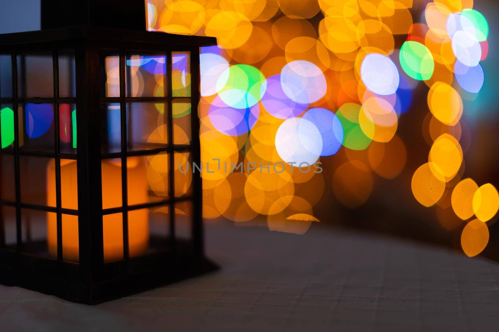 In the night frame a rectangular old hand lantern with a glowing candle in the center. In the distance a blurred background created from the bokeh of glowing Christmas tree lights