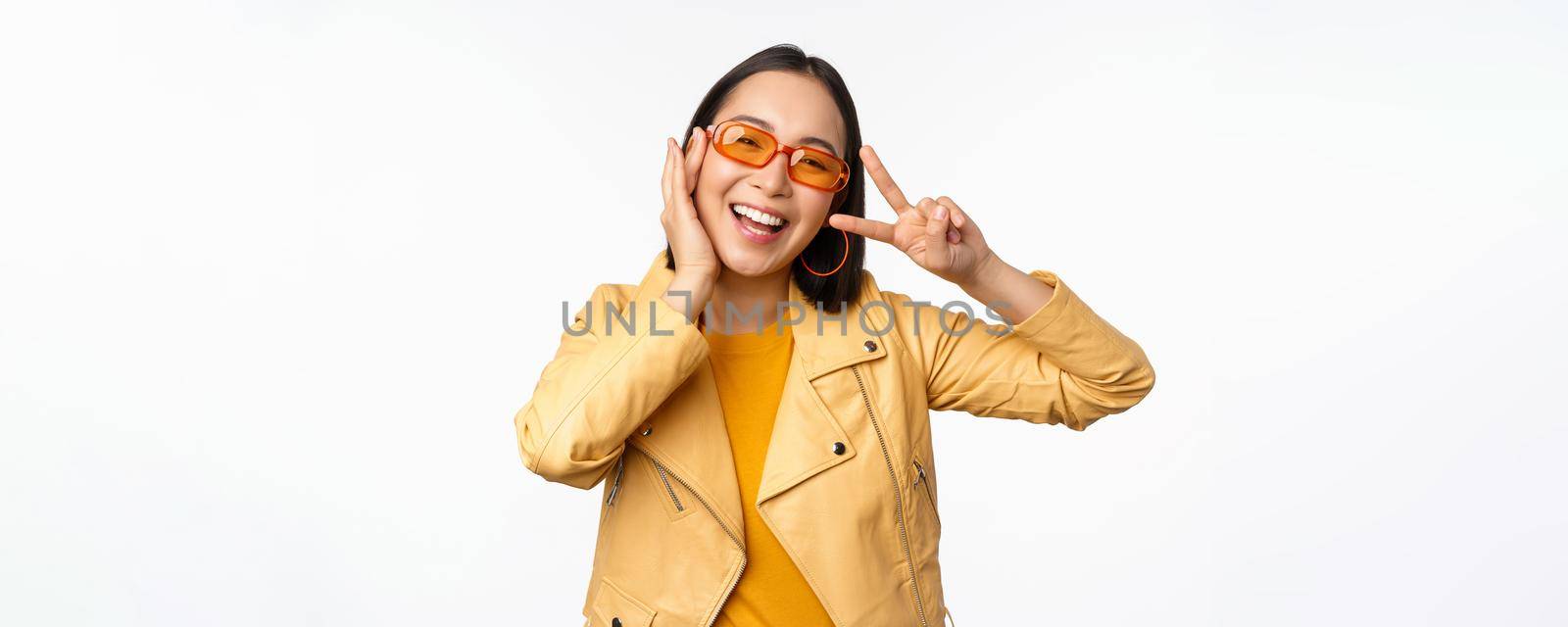 Portrait of stylish asian modern girl, wearing sunglasses and yellow jacket, showing peace, v-sign gesture, standing over white background, happy smiling face.