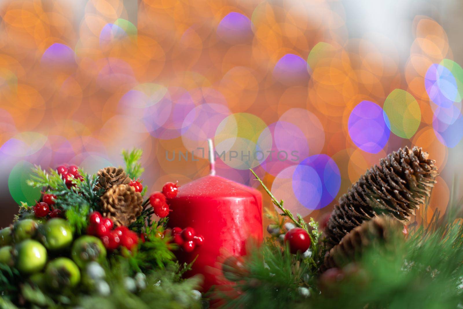 A close up shot shows a red candle sticking out of a Christmas wreath. In the distance a blurred background created from the bokeh of the glowing Christmas tree lights