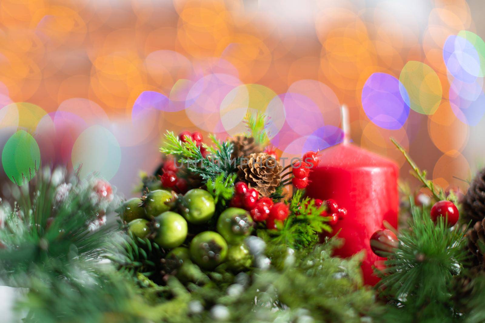 A close up shot shows a red candle sticking out of a Christmas wreath. In the distance a blurred background created from the bokeh of the glowing Christmas tree lights