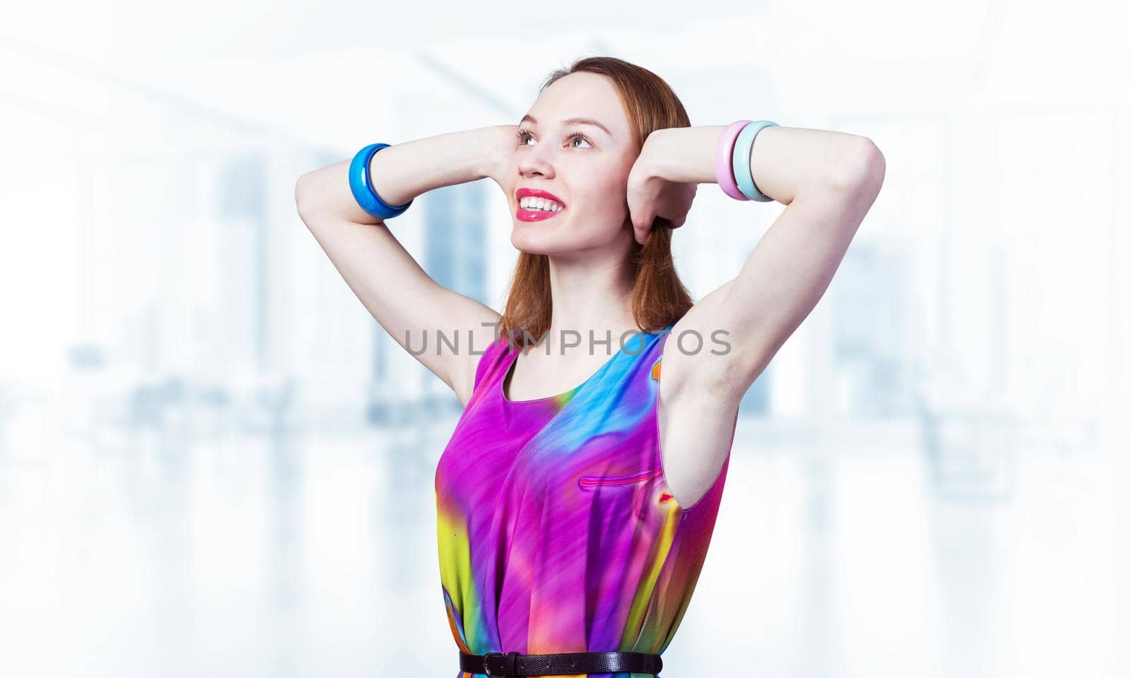 Redhead beautiful girl closed her ears with hands. Joyful lady with charming smile in bright colorful dress and bracelets on blurred office background. Portrait of happy young woman looking up aside.