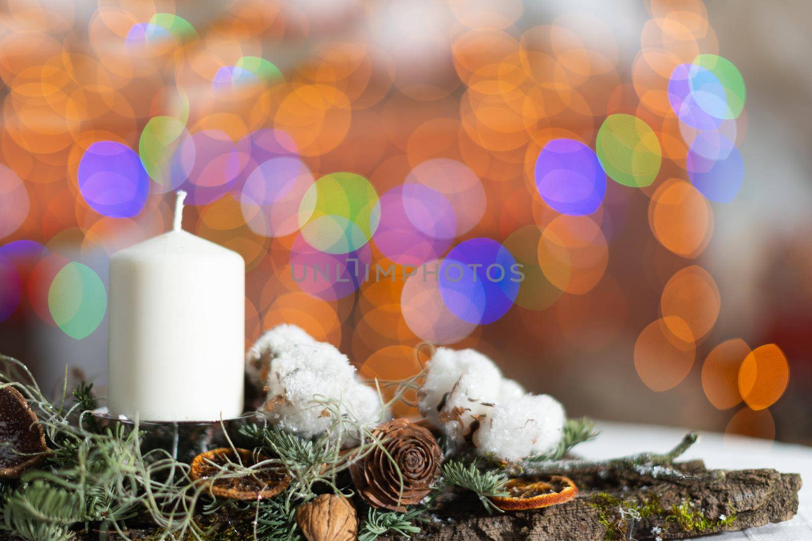 In the close up frame you can see a white candle sticking out of a Christmas wreath. In the distance a blurred background created from the bokeh of the glowing Christmas tree lights