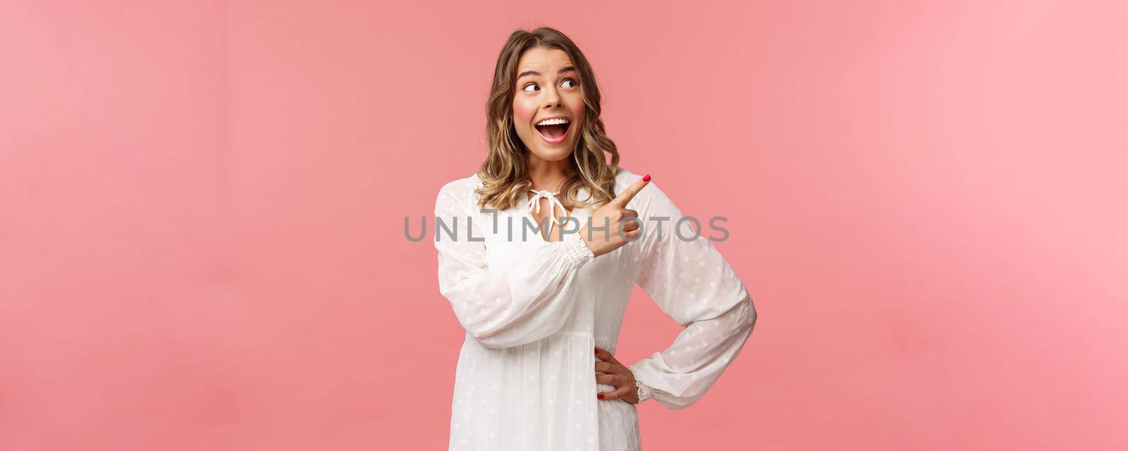 Enthusiastic, happy attractive blond woman in white cute dress, open mouth excited and cheerful looking, pointing upper right corner as best prices discounts ever, stand pink background by Benzoix